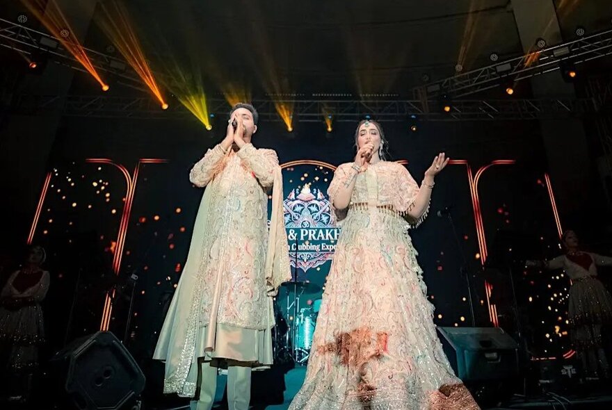 Two Bhajan performers in glittering traditional clothes singing on stage.