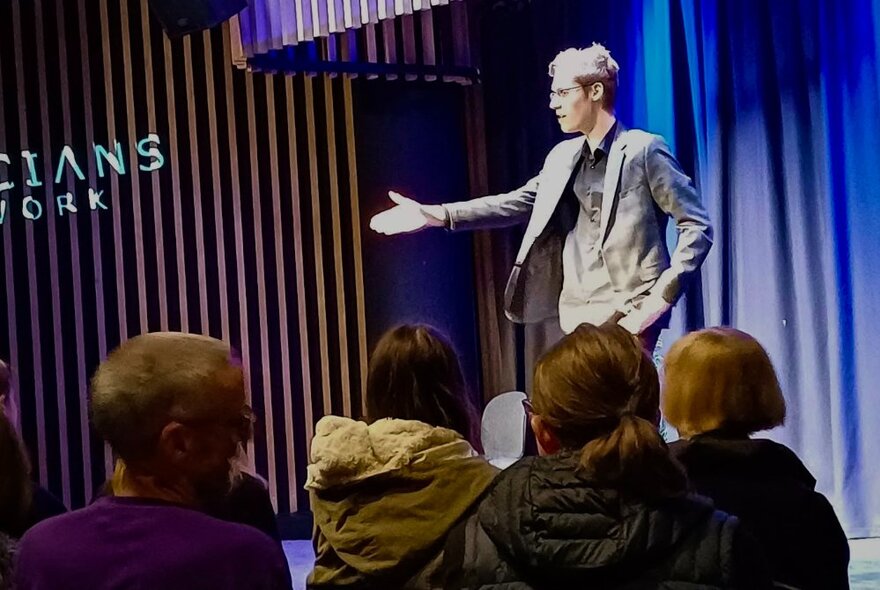 Performer on stage with one arm raised, other hand in pocket, audience looking on.
