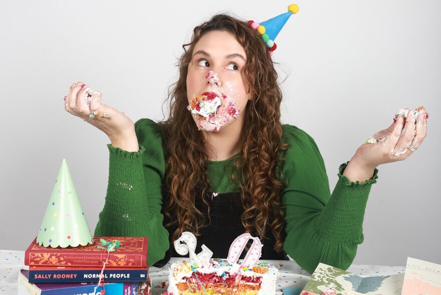 Performer Abby Hampton wearing a party hat, her face, mouth and hands covered in cake, resting her elbows on a table in front of her.