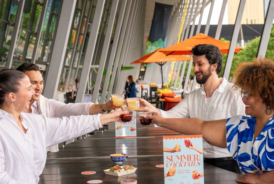 Four people seated at an outdoor table at MCEC, clinking their glasses together above the table. Orange umbrellas in the background.