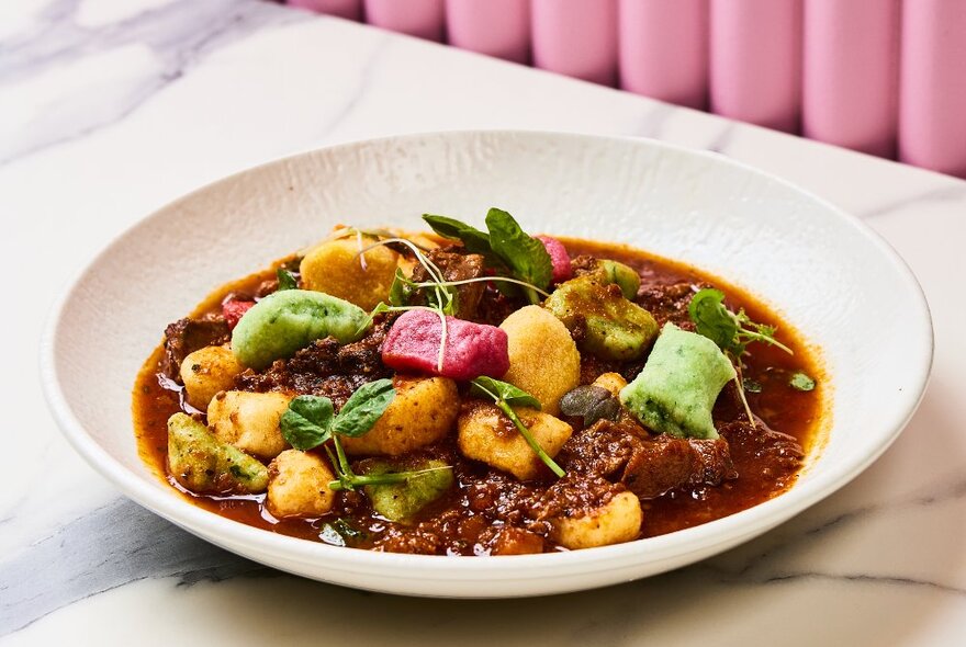 A bowl of green, pink and yellow gnocchi in a bowl of brown sauce on a marble table.