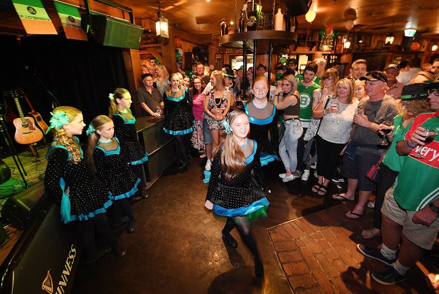 A lively St. Patrick's Day celebration at a crowded Irish pub in Melbourne, featuring patrons dressed in festive green attire watching girls perform traditional Irish dancing.