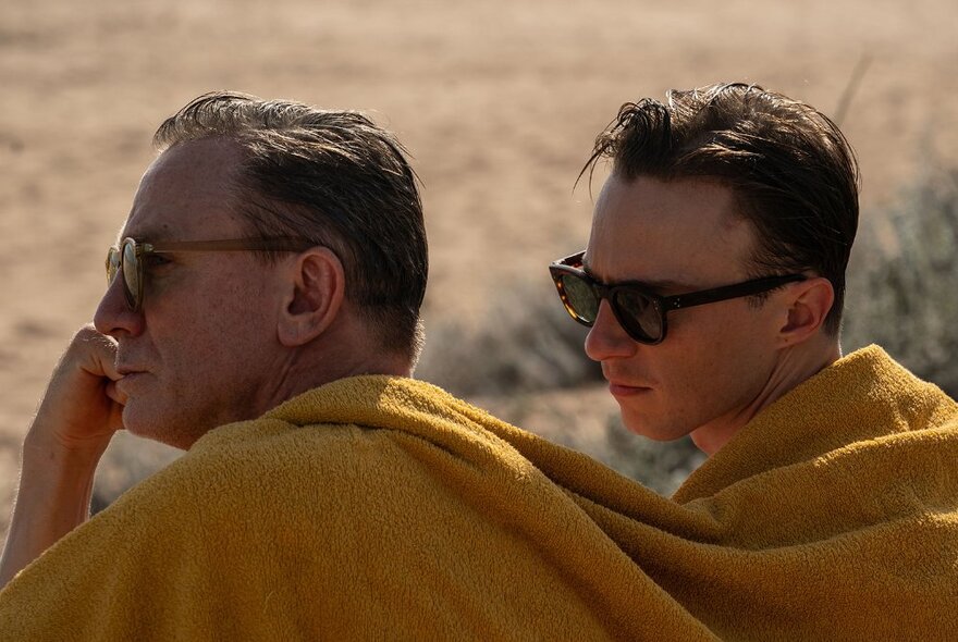 Film still of two men in profile, sitting at a sandy place outdoors, their shoulders covered by a yellow blanket or large towel, both wearing sunglasses.