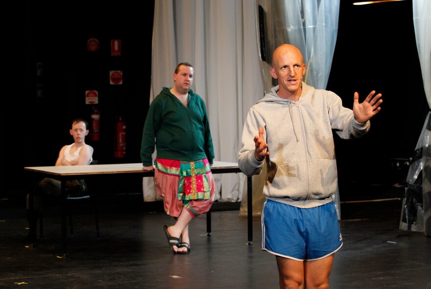 Three people performing on stage, the person at the front in blue running shorts and a tracksuit top making gestures with his hands. 