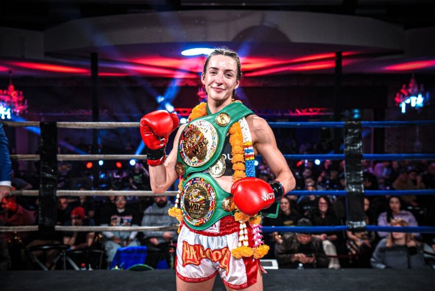 A boxing champion dressed in boxing belt medals with her gloves still on in the ring. 