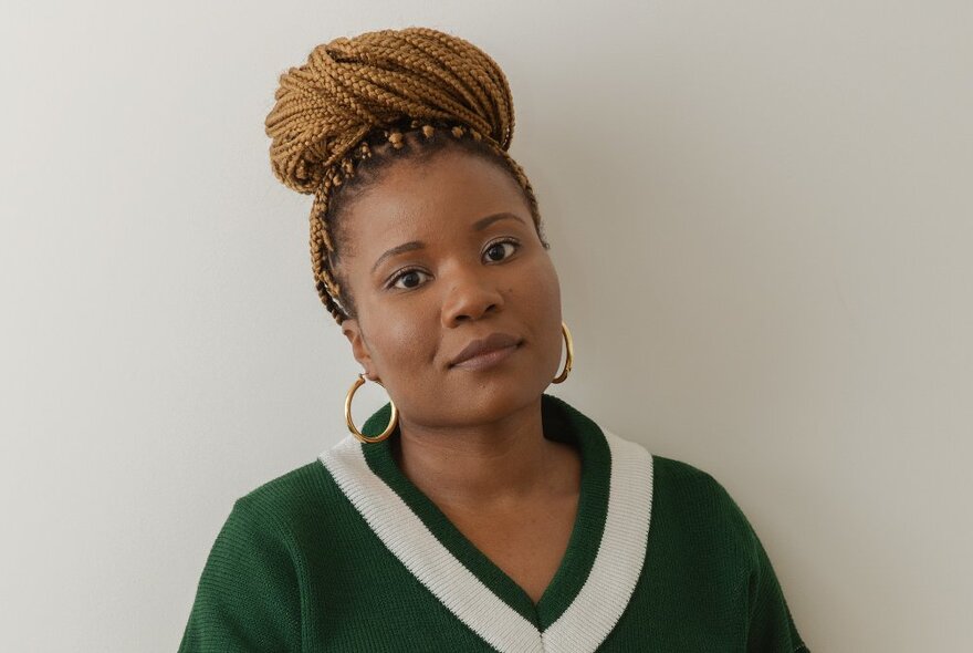 Author Santilla Chingaipe posed against a white wall, with her long braided hair curled into a large bun on top of her head, wearing gold hoop earrings and a V-necked top.