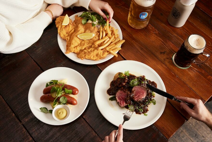 Person's hand taking a chip from a plate of fish and chips, other person's hands holding knife and fork over a dish of meat, on a restaurant table with another dish of sausages and two steins of beer.