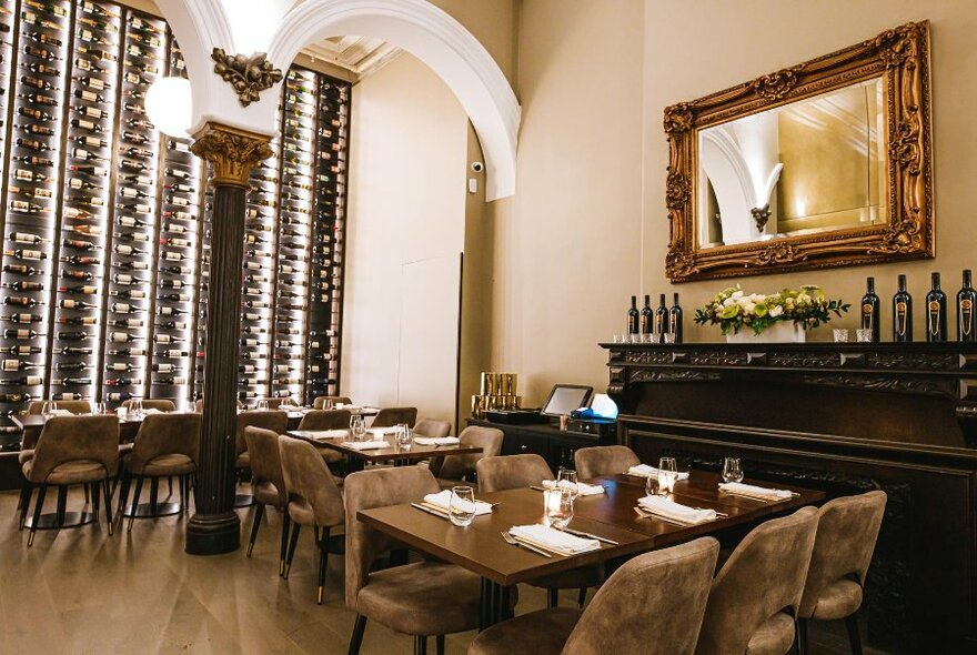 Interior of a restaurant with tables and chairs set for service, ornate ceiling details, a wall of wines and a large mirror in a gilded frame resting on a wall above a mantelpiece.