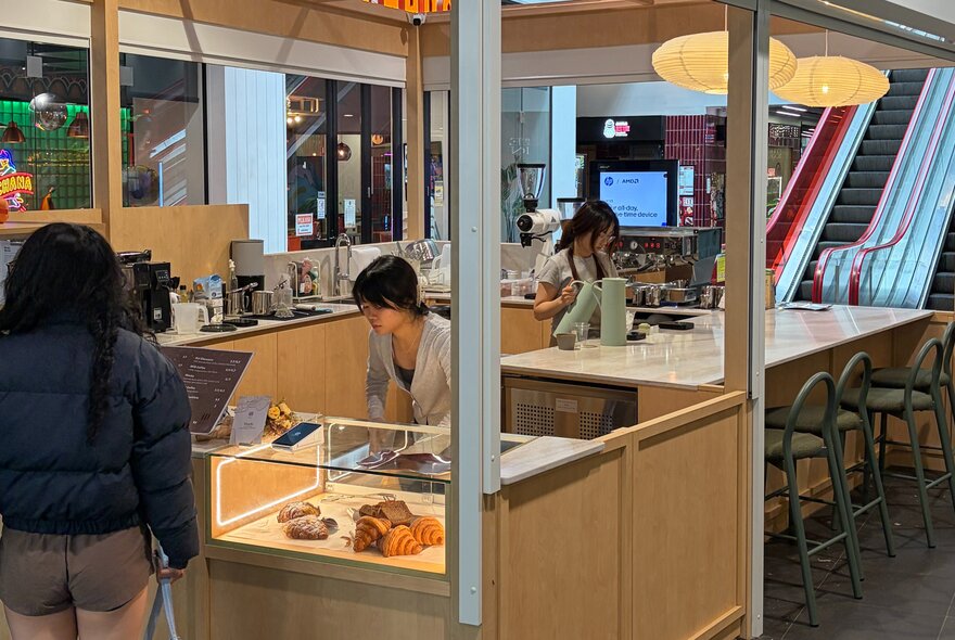 A pale timber booth in a shopping centre serving pastries and coffee, staff serving at the counter. 