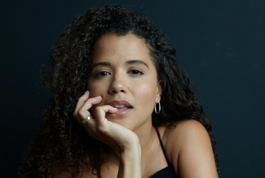 Playwright Kirsty Marillier, a young woman with long curly dark hair framing her face, her chin resting in her hand, with her little finger tip resting on her bottom lip. 