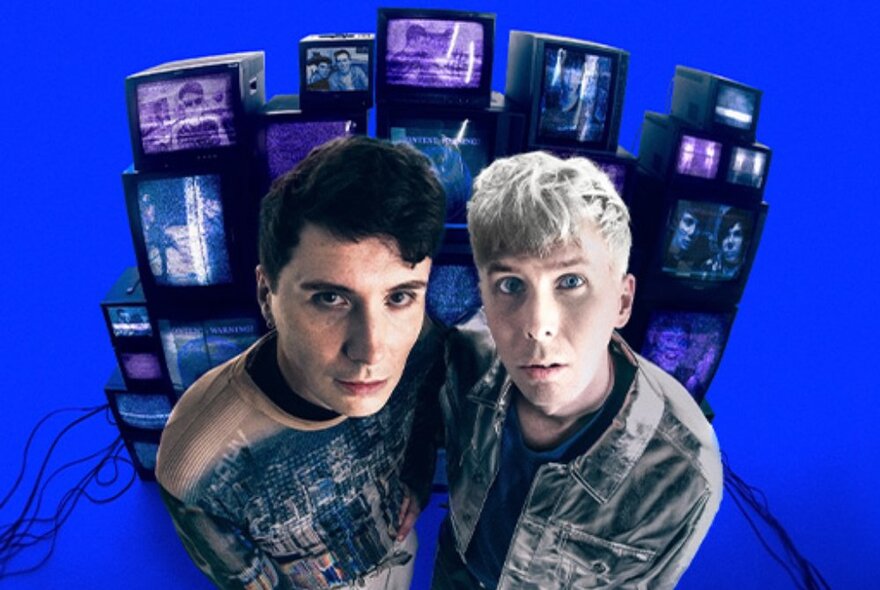 Comedians Dan and Phil standing side by side in front of a stack of television screens; image taken with a fisheye lens.