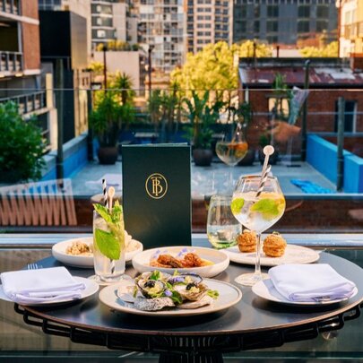 A rooftop table set with food and drinks on a glass-bordered terrace overlooking nearby buildings.
