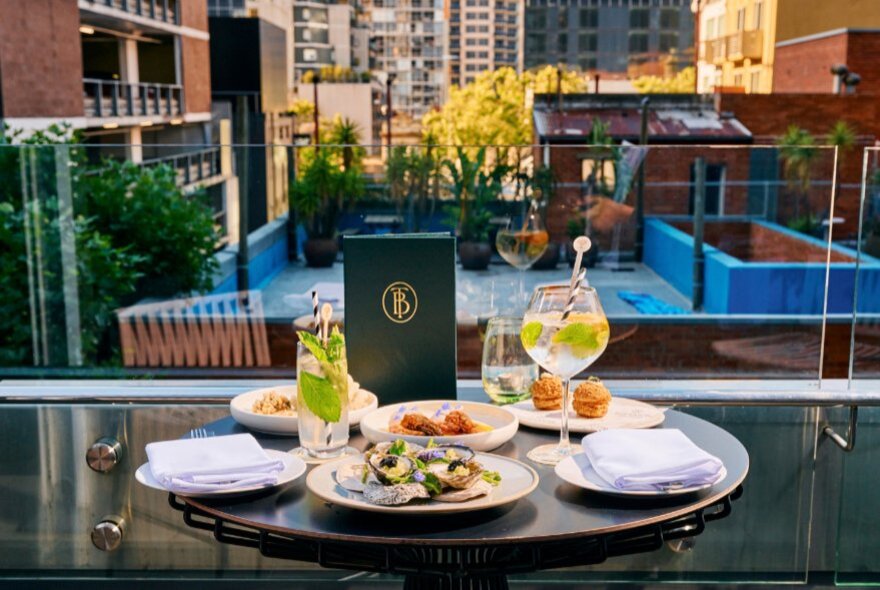 A rooftop table set with food and drinks on a glass-bordered terrace overlooking nearby buildings.