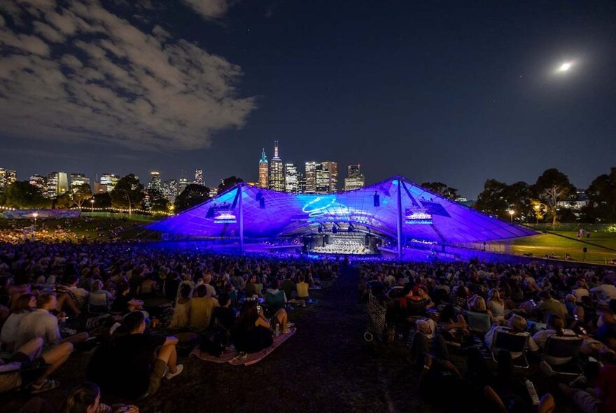 A large crowd seated on the lawns surrounding the Sidney Myer Music Bowl at night, with the venue in mid-distance illuminated in blue lights, and the outline of city buildings visible in the far background.