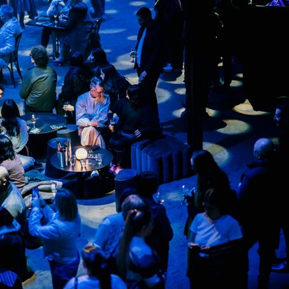 Visitors enjoying an evening event at the National Gallery of Victoria, seated on stools under shadowy lighting.