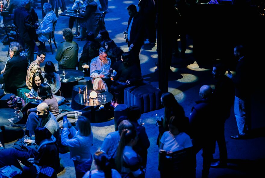 Visitors enjoying an evening event at the National Gallery of Victoria, seated on stools under shadowy lighting.
