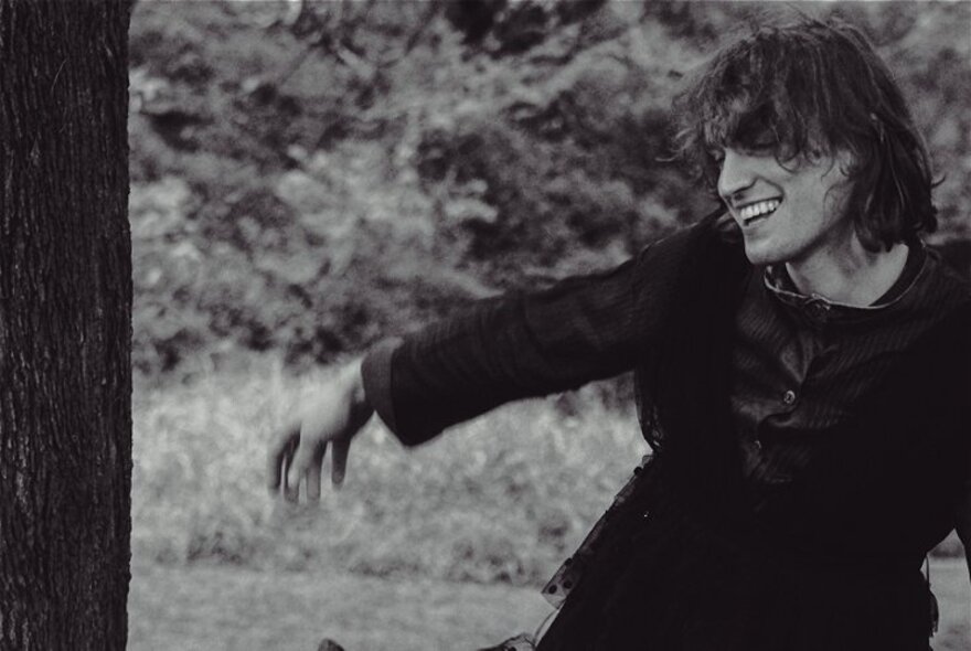 A black and white image of a smiling man wearing black, with one arm outstretched.