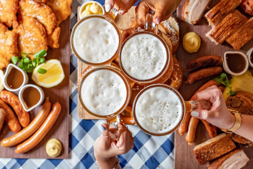 Looking down from above with hands holding four large steins of beer over a table filled with German style food platters. 