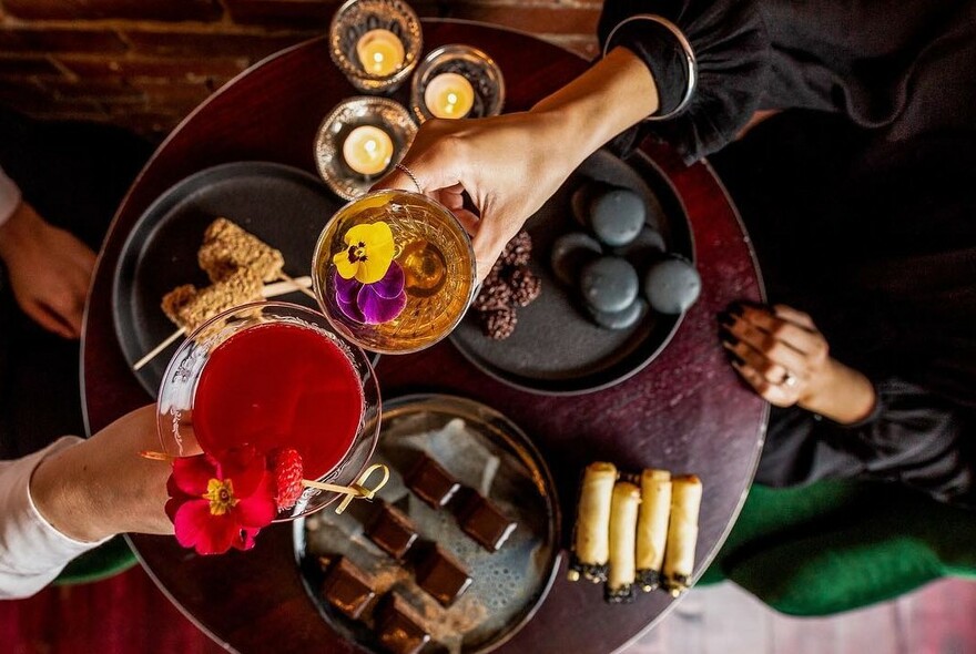 People clinking cocktail glasses over a small table with snacks.