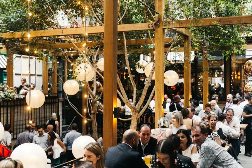 A busy beer garden with trees and hanging lanterns