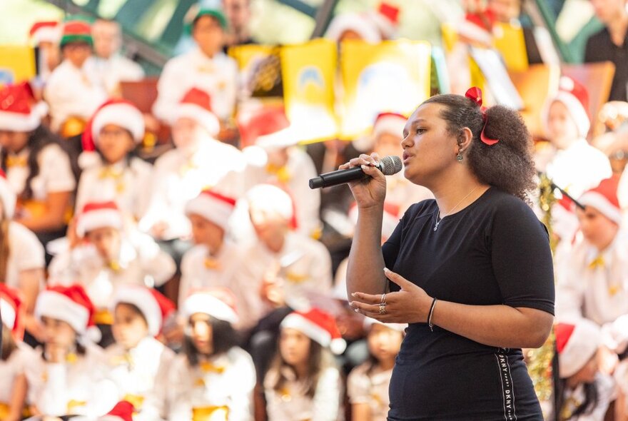 A soloist singing into a microphone in the foreground, with a choir of children behind her wearing festive red Christmas hats.