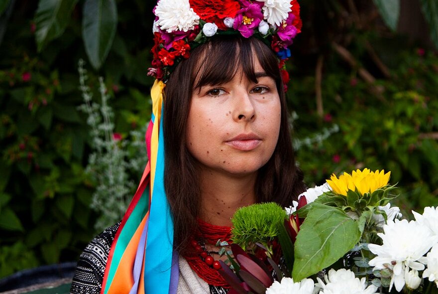 A woman staring at the viewer, holding a large bunch of flowers, wearing a brightly coloured floral headpiece with long ribbons hanging from it.