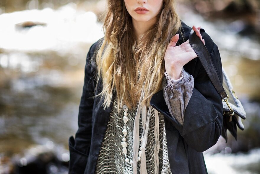 A woman with the top part of her face chopped off waring a drapy knit under a jacket and a holding a bag over her shoulder with her palm exposed. 