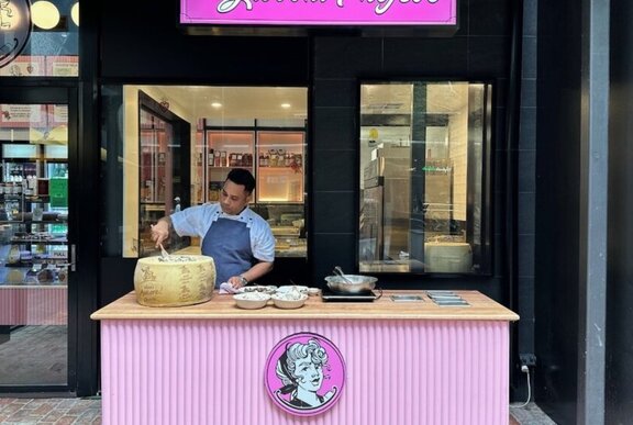 Man standing in front of a table, twirling pasta in a large parmesan wheen to dish up into bowls, in front of a restaurant.