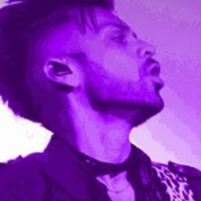 A purple-washed profile headshot of a Prince impersonator singing. 