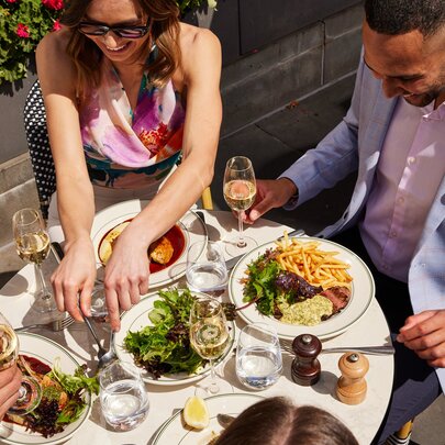 An outdoor restaurant table with plates of food and glasses of wine, with three people around it eating and interacting with each other.