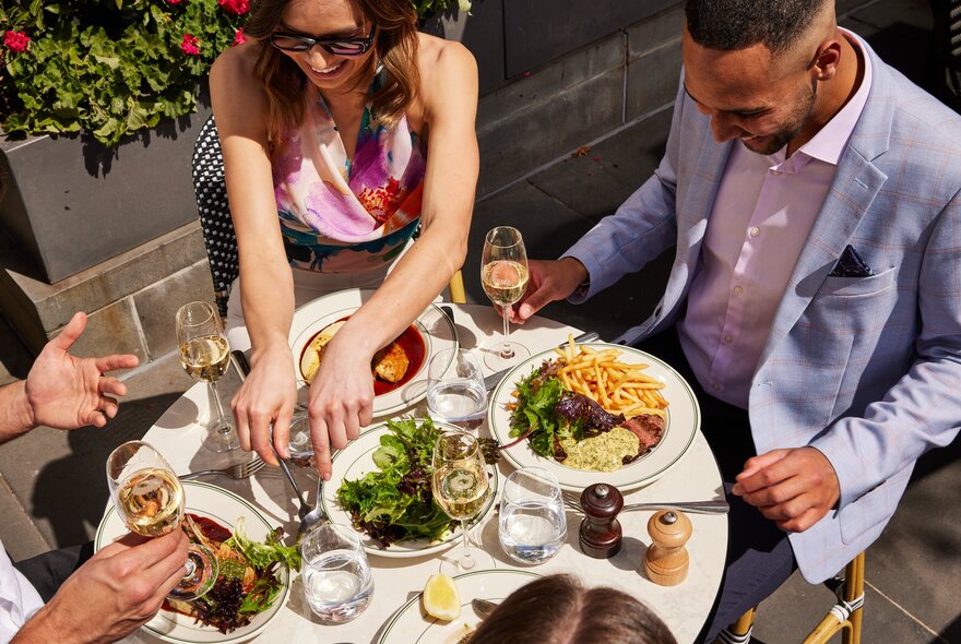 An outdoor restaurant table with plates of food and glasses of wine, with three people around it eating and interacting with each other.