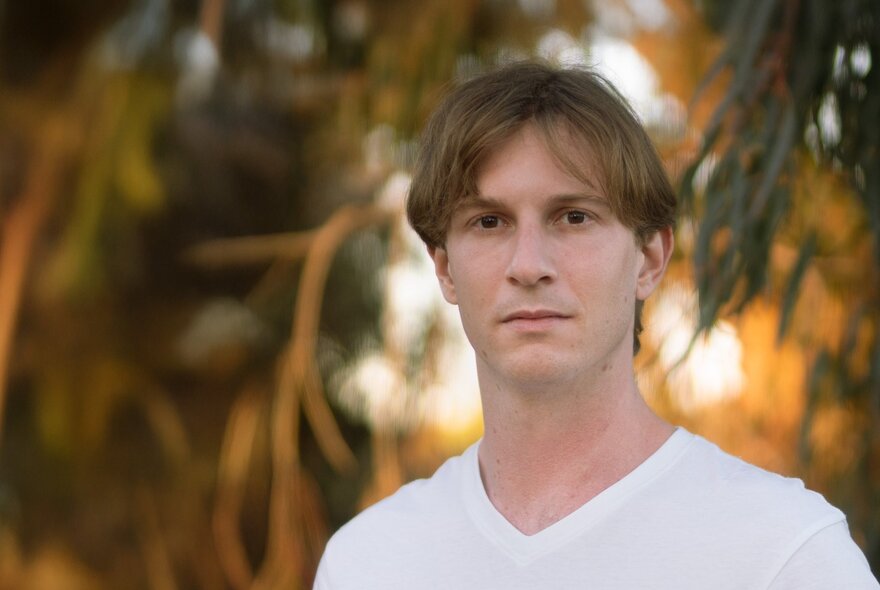 A man wearing v-necked white tshirt looking blank in a garden setting with sunlight glowing through a tree behind him. 