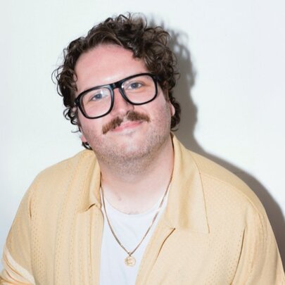 Australian comedian and film critic Alexei Toliopoulos; he wears a pale shirt and glasses, has a moustache and brown wavy hair and is smiling at the camera.