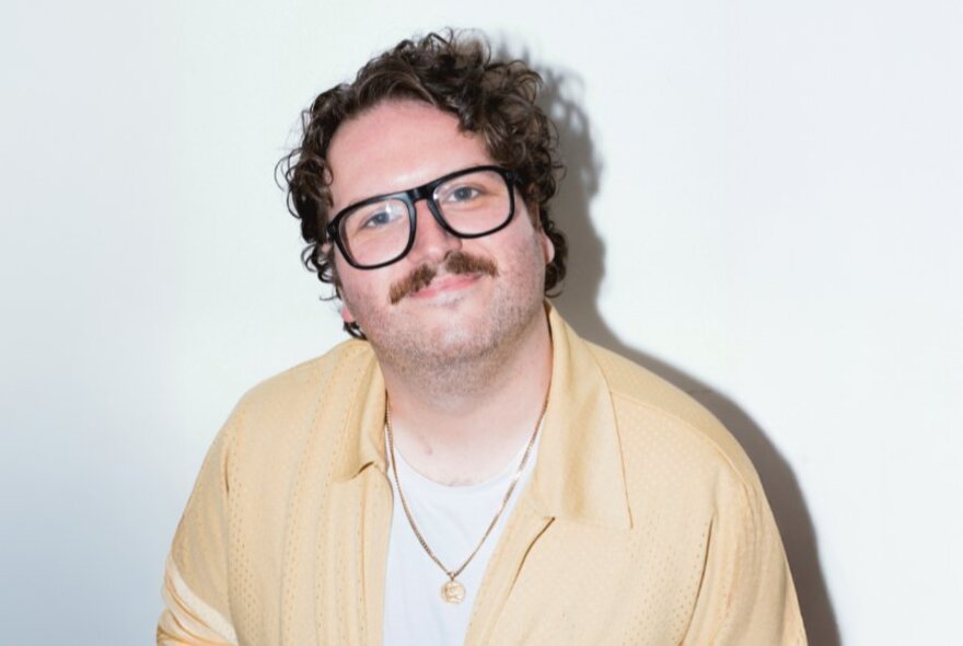 Australian comedian and film critic Alexei Toliopoulos; he wears a pale shirt and glasses, has a moustache and brown wavy hair and is smiling at the camera.
