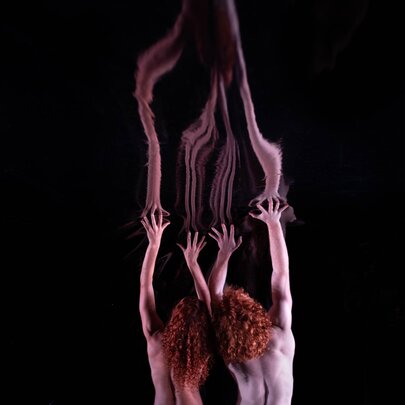 A scene from the contemporary dance work Red by Australian company Dancenorth, featuring two people from the back, with their heads close together and their arms reaching upward into a dark void.