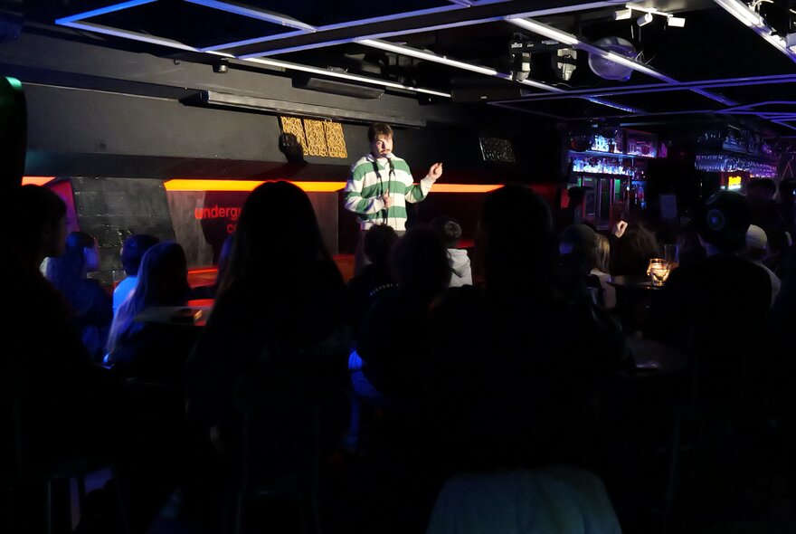 Stand-up comedian standing on a stage holding a microphone in front of a small seated audience.
