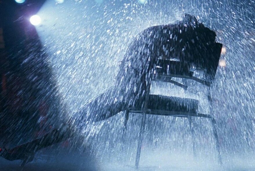 A scene from the film Flashdance with a dancer in silhouette leaning back in a chair, a spotlight behind them, surrounded by water droplets.