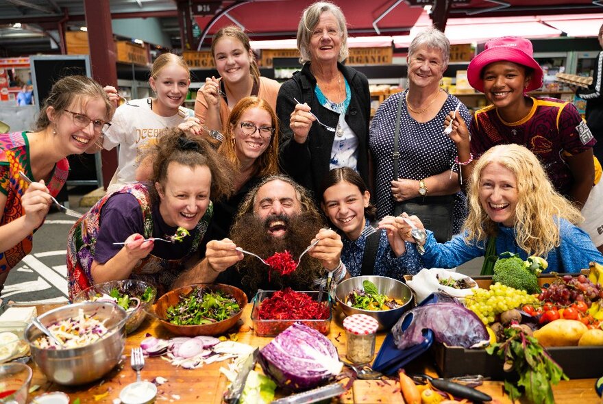 Celebrity gardener Costa being fed red food at a large communal table, surrounded by smiling people with forks.