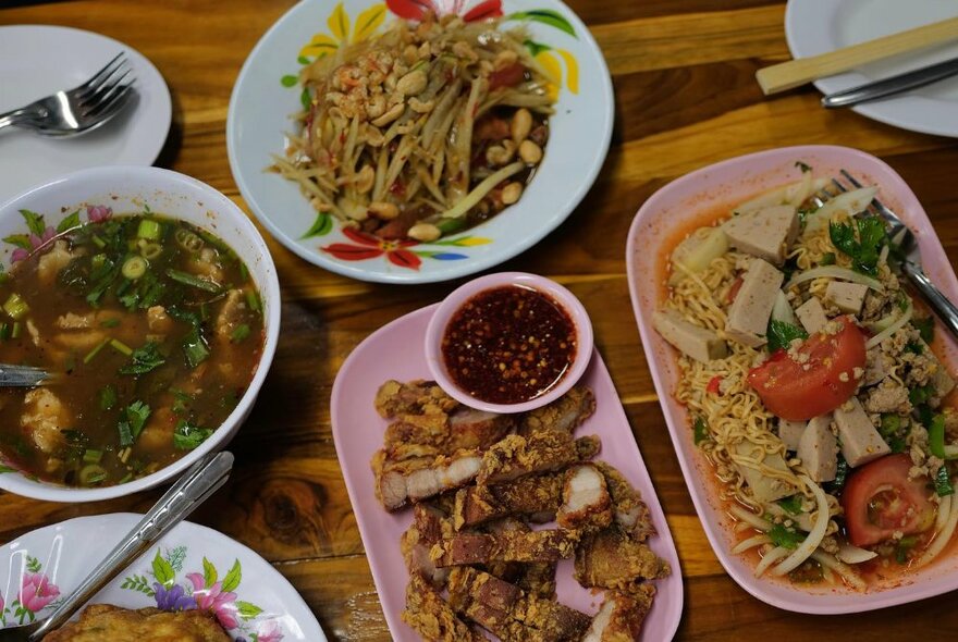 Several Thai dishes on a cafe table with cutlery and chopsticks including noodles, rice and soup.