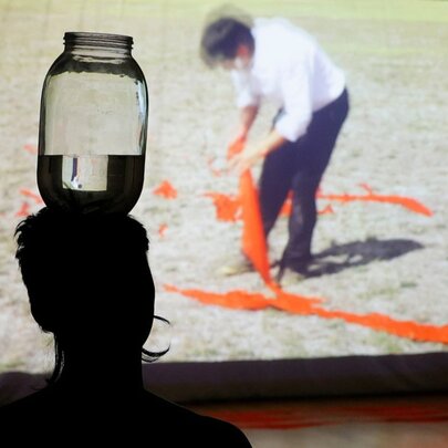 A silhouette of a person balancing a jar of water on their head while watching a large screen showing an a man with orange streamers. 