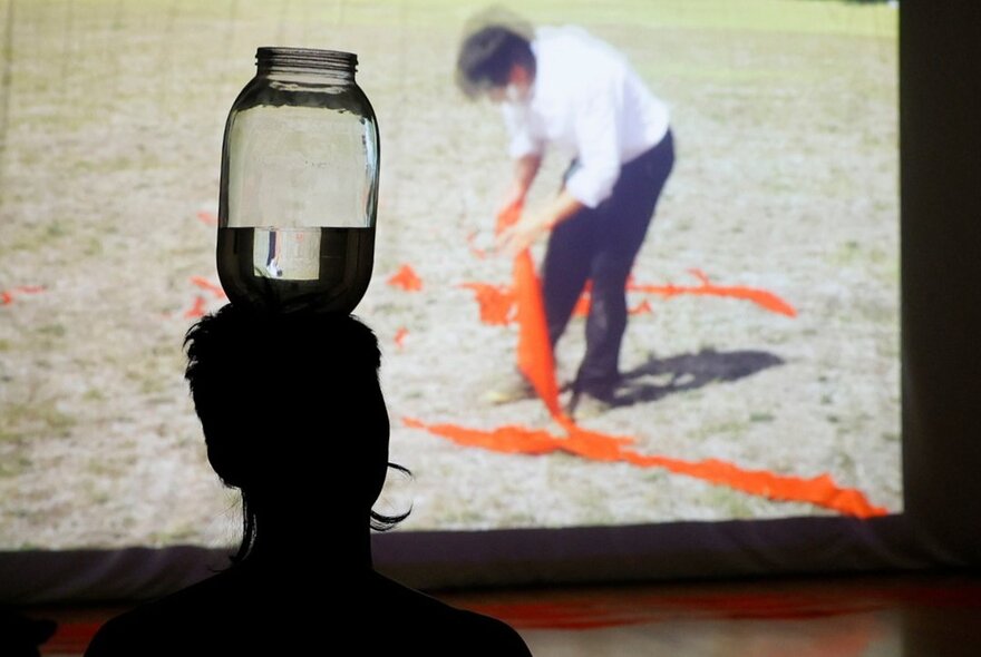 A silhouette of a person balancing a jar of water on their head while watching a large screen showing an a man with orange streamers. 