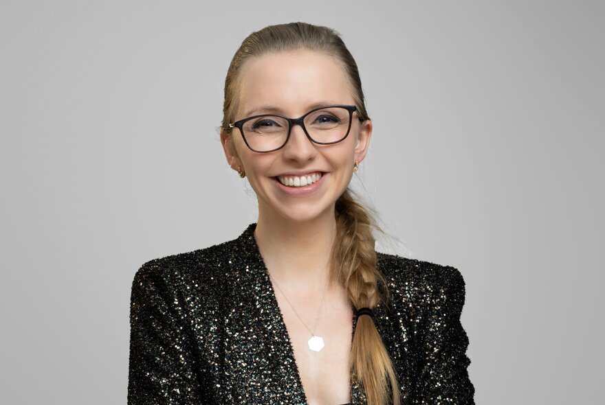 A portrait photo of Anna Lapwood, a woman with blonde hair in a side braid, wearing glasses and a black sequined jacket, smiling at the camera. 