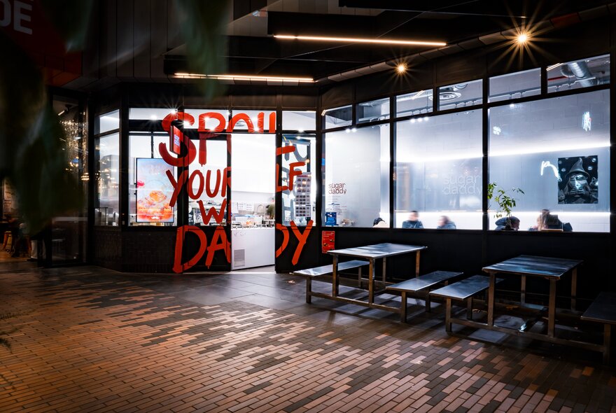 The exterior of a dessert bar at night with some benches out the front and red writing splashed over the window and door. 