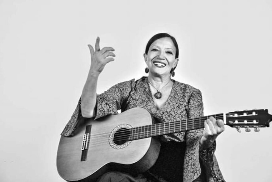 Black and white photograph of smiling Italian singer, Sara Cappella, with an acoustic guitar and one hand up in the air.