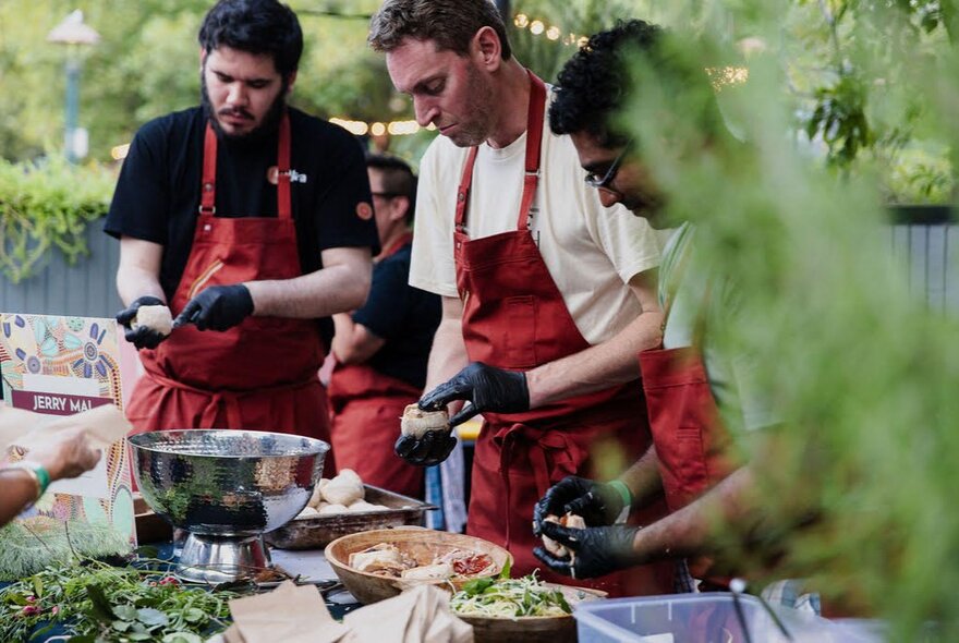 A group of men wearing matching maroon aprons, all preparing food. 