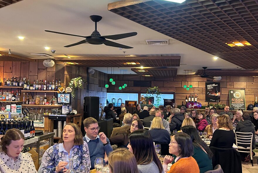 A busy dining area with people interacting at tables and a large ceiling fan with black blades. 