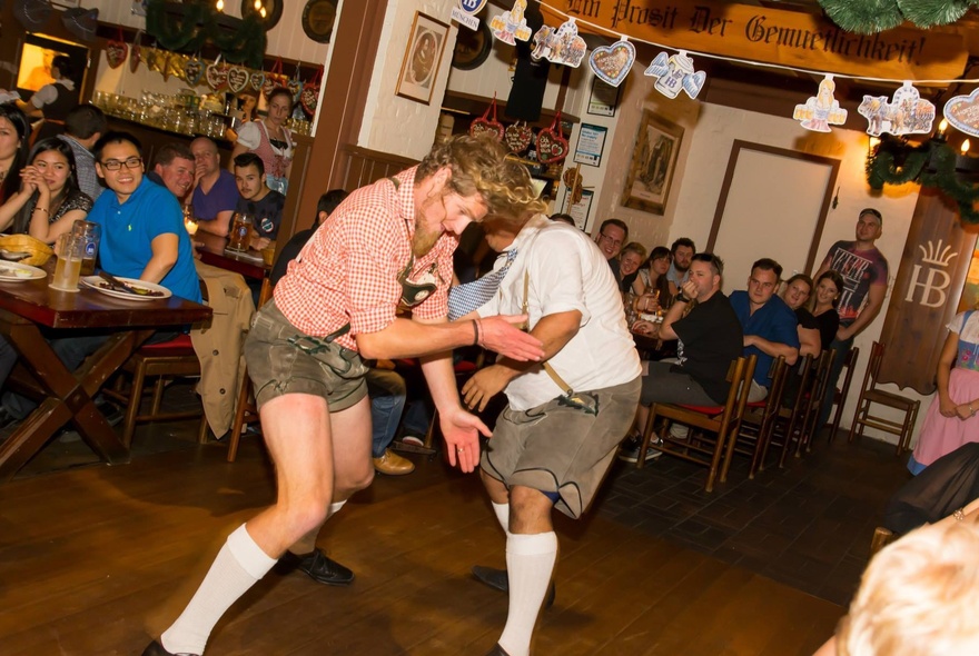 Two men wearing Tyrolean shorts and socks, wrestle-dancing in a restaurant.