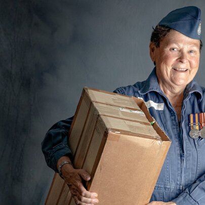 A smiling woman in an Australian military uniform, adorned with medals, holding a large cardboard box.