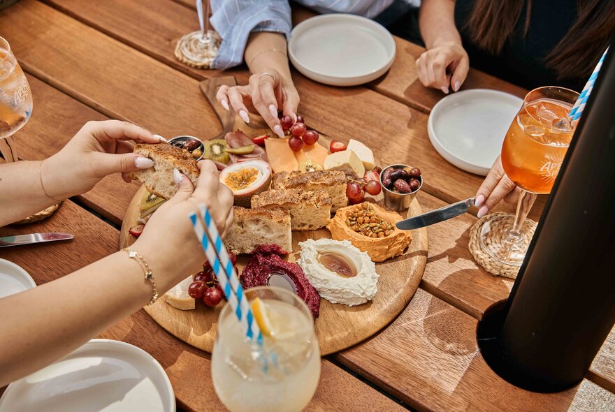 Hands hovering over an antipasto wooden platter on a table, with drinks and small side plates in front of them.