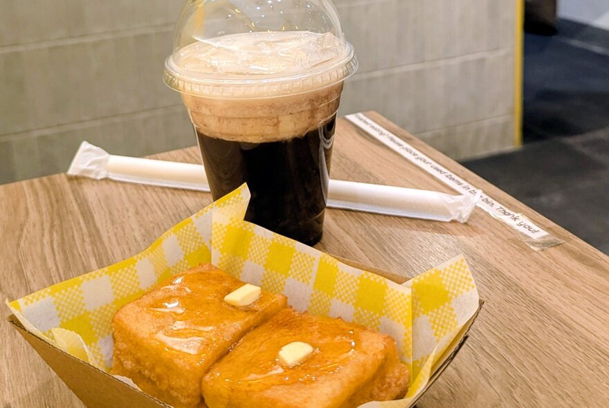 Hong Kong-style French Toast served in a cardboard tray with yellow paper lining, on a table next to an iced coffee.